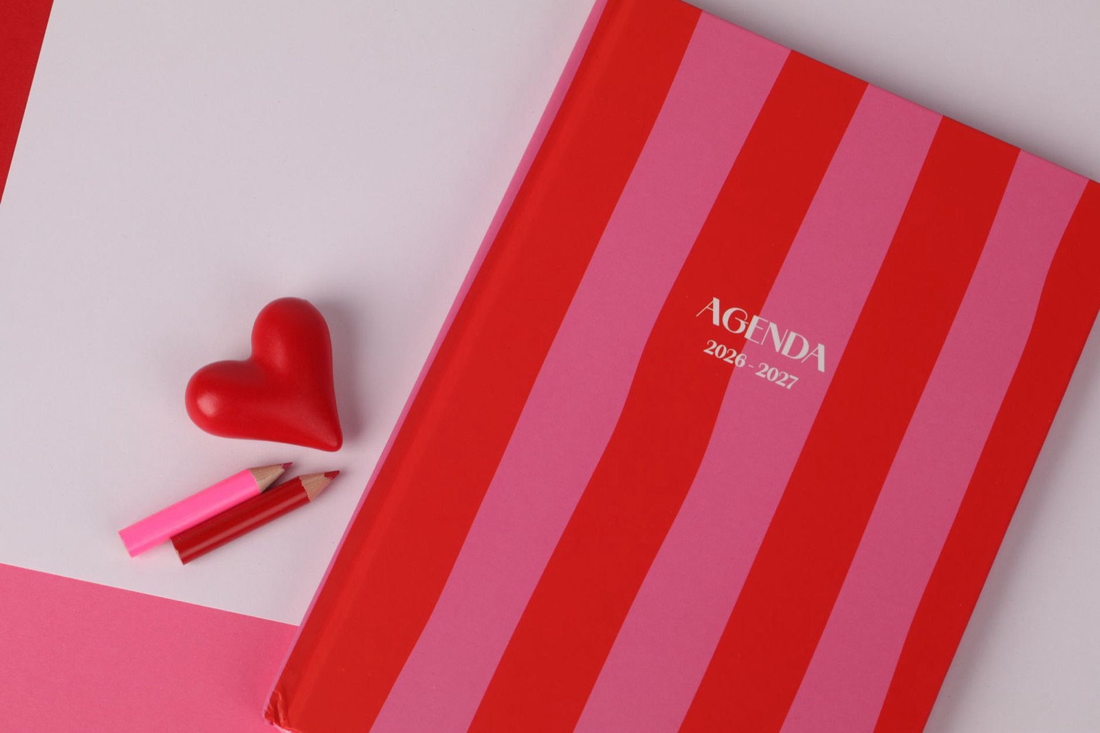 Flat lay of a red and pink striped agenda, heart-shaped object, and colored pencils, ideal for planning themes.