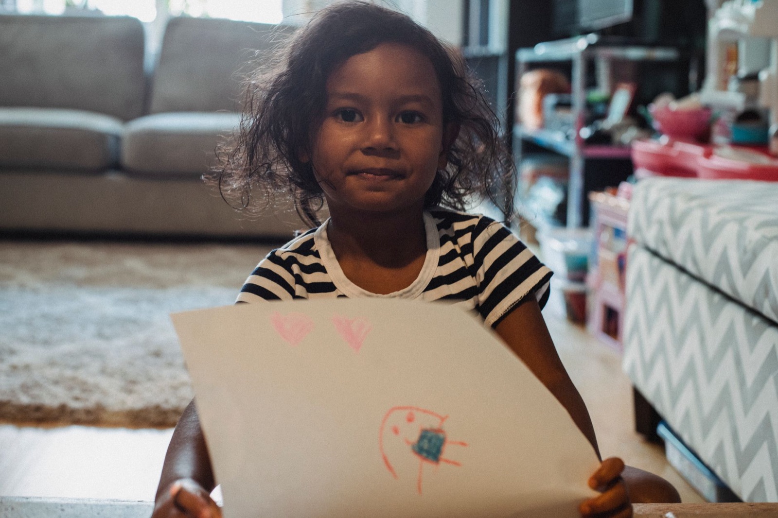 Cute ethnic girl in casual shirt demonstrating paper sheet with drawing while sitting in living room and looking at camera
