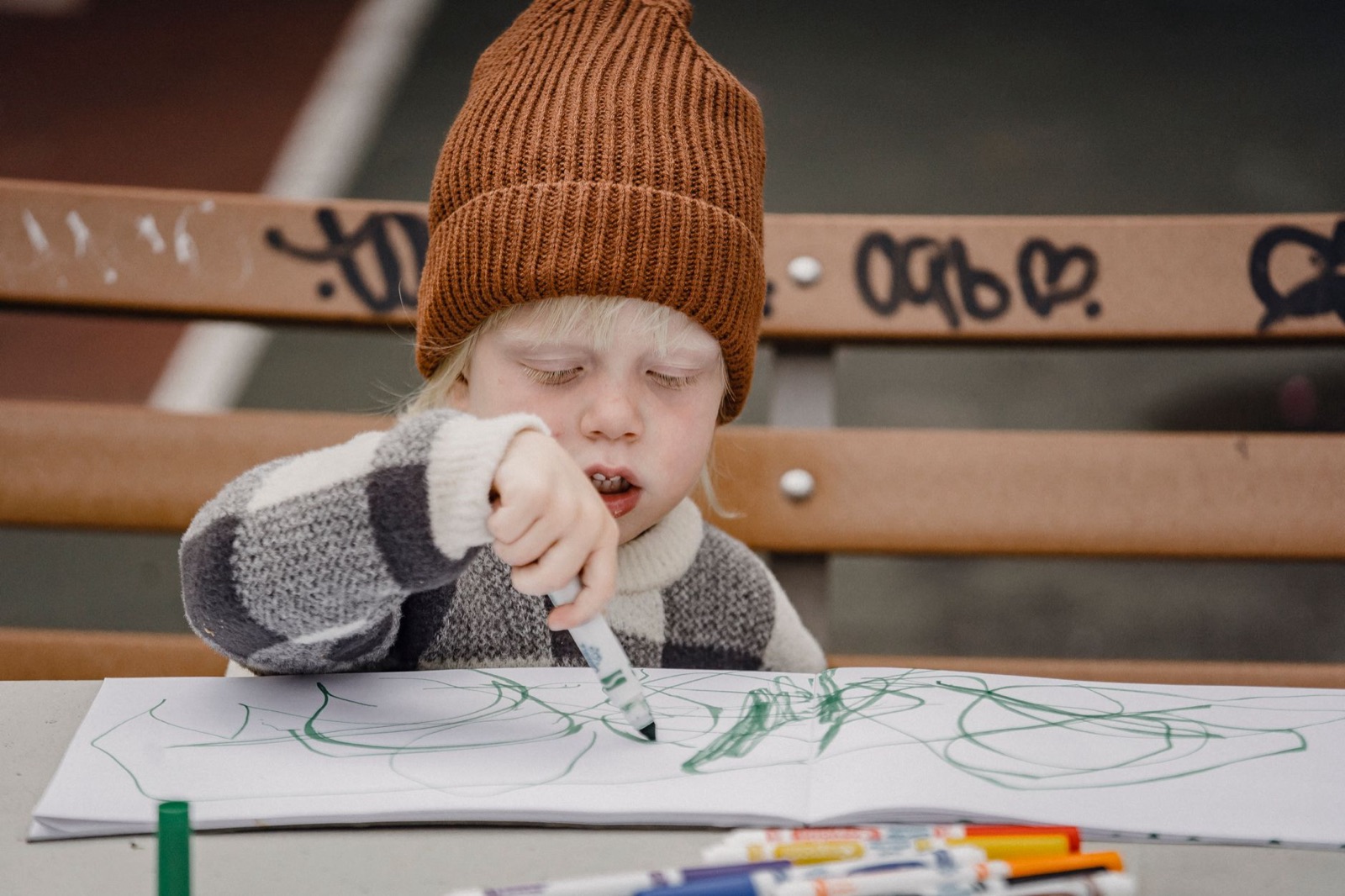 Focused little lid wearing hat drawing with green marker on paper while sitting on wooden bench at table on street against blurred background
