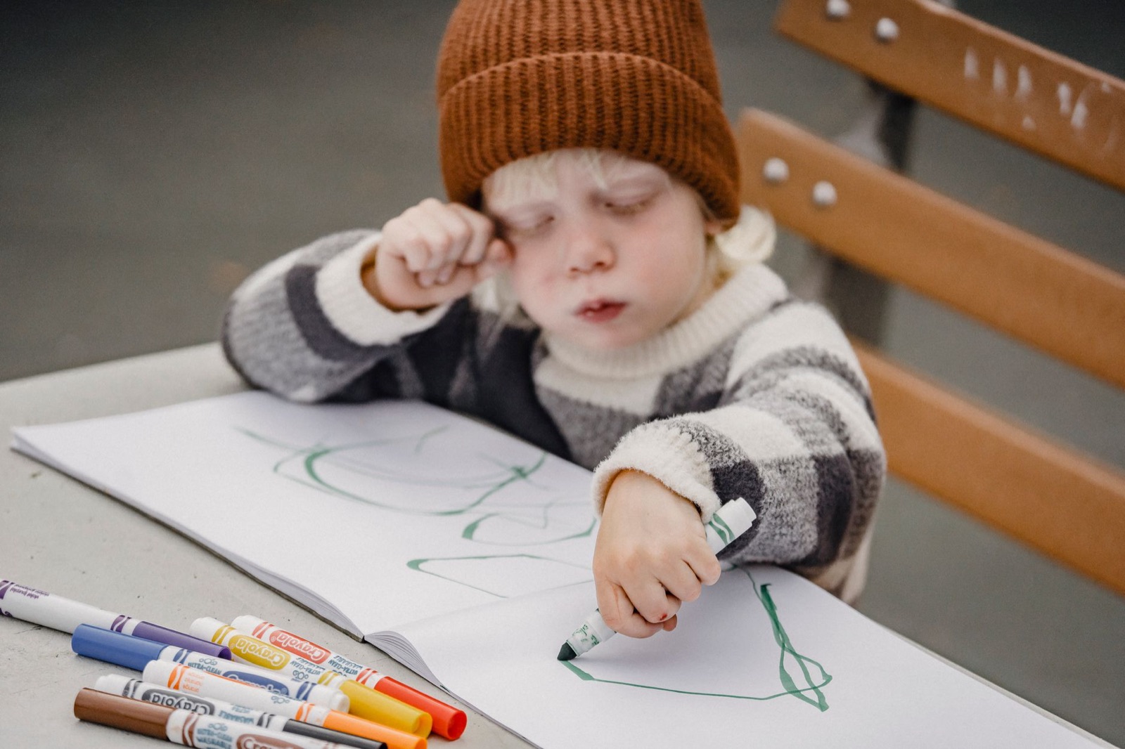 Crop focused little kid wearing hat sitting on wooden bench at table while drawing with green felt pen on white paper against blurred background