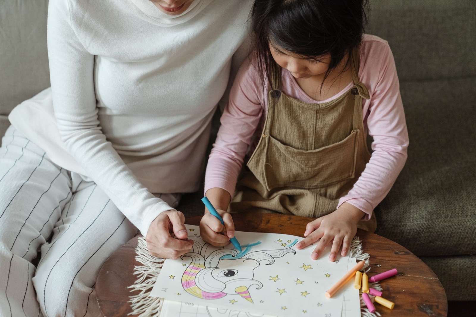 From above crop concentrated Asian girl wearing casual clothes and sitting on cozy sofa with mother while coloring picture on paper with crayons of different color