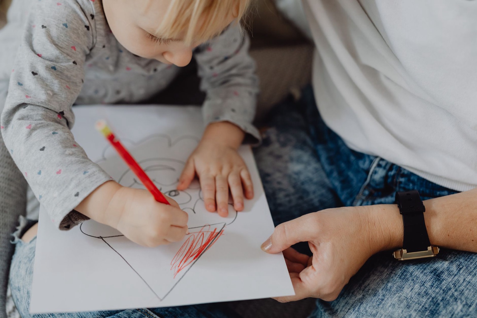 A young child focused on coloring a drawing with a red pencil under adult supervision.