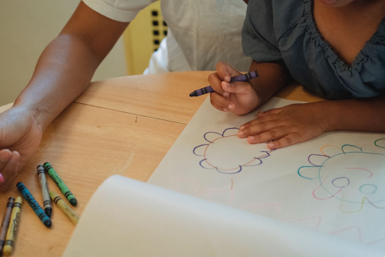 Crop anonymous mother and daughter sitting at wooden table and drawing with color wax crayons