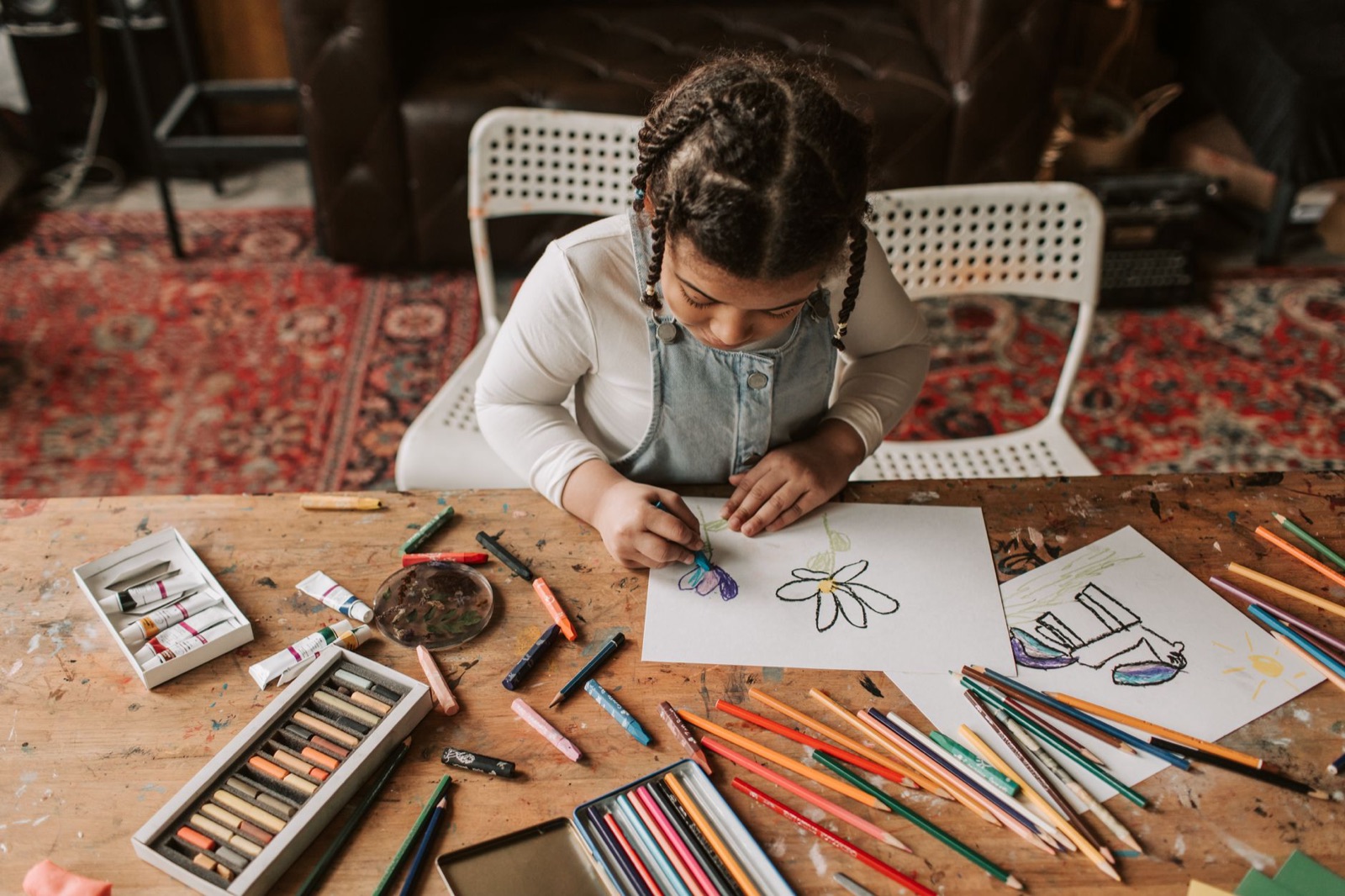 A young girl focused on drawing with crayons and colored pencils at a wooden table indoors.