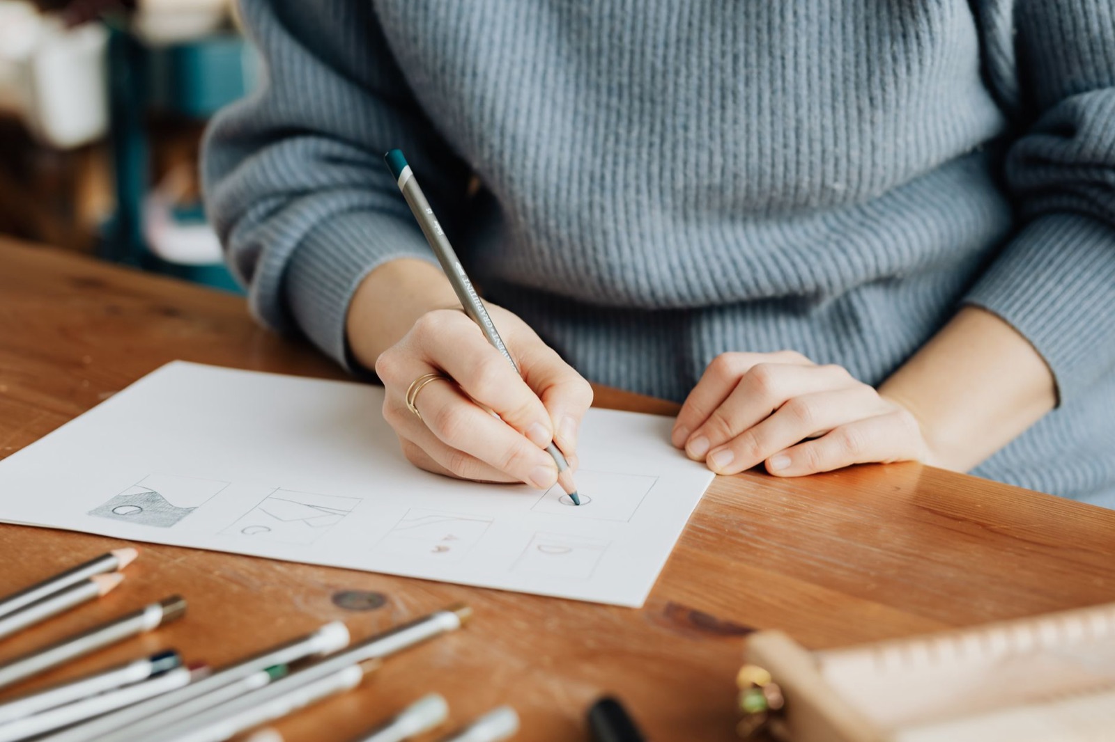Crop anonymous female illustrator in knitted sweater using pencil while drawing on sheet of paper and sitting at wooden desk in light office