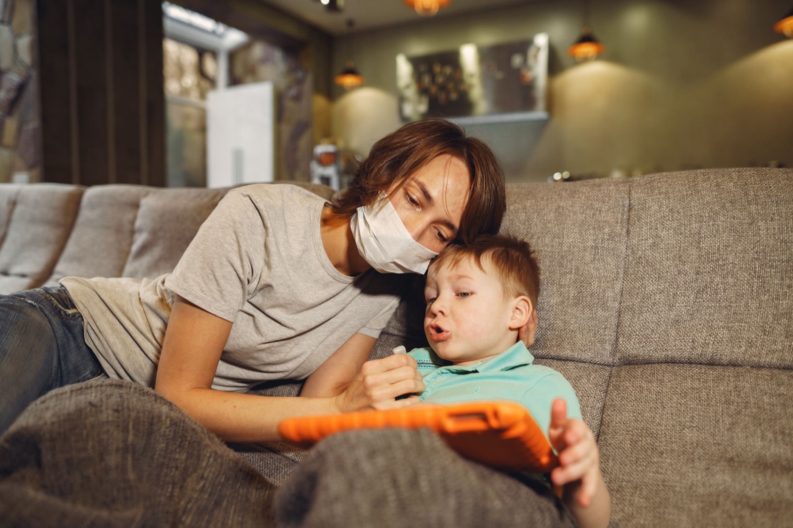 Mother in face mask sitting on cozy couch with little boy covered with blanket while watching tablet and talking in apartment