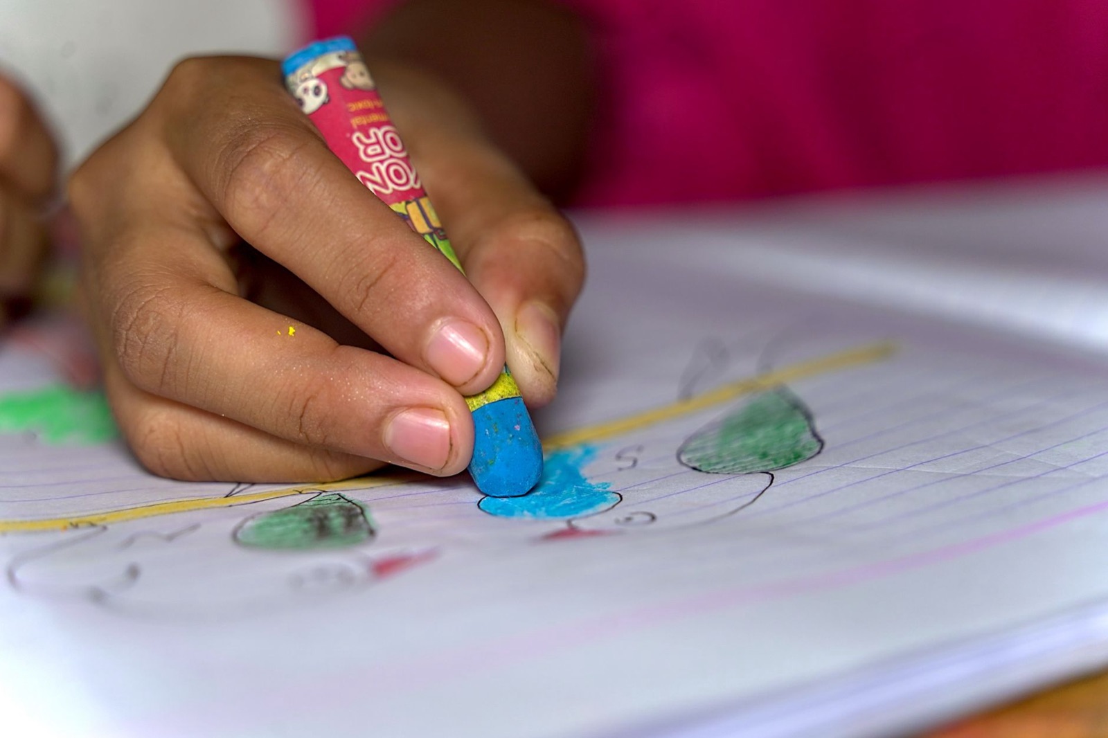 Uma criança colorindo um desenho com um giz de cera azul em uma folha de caderno.