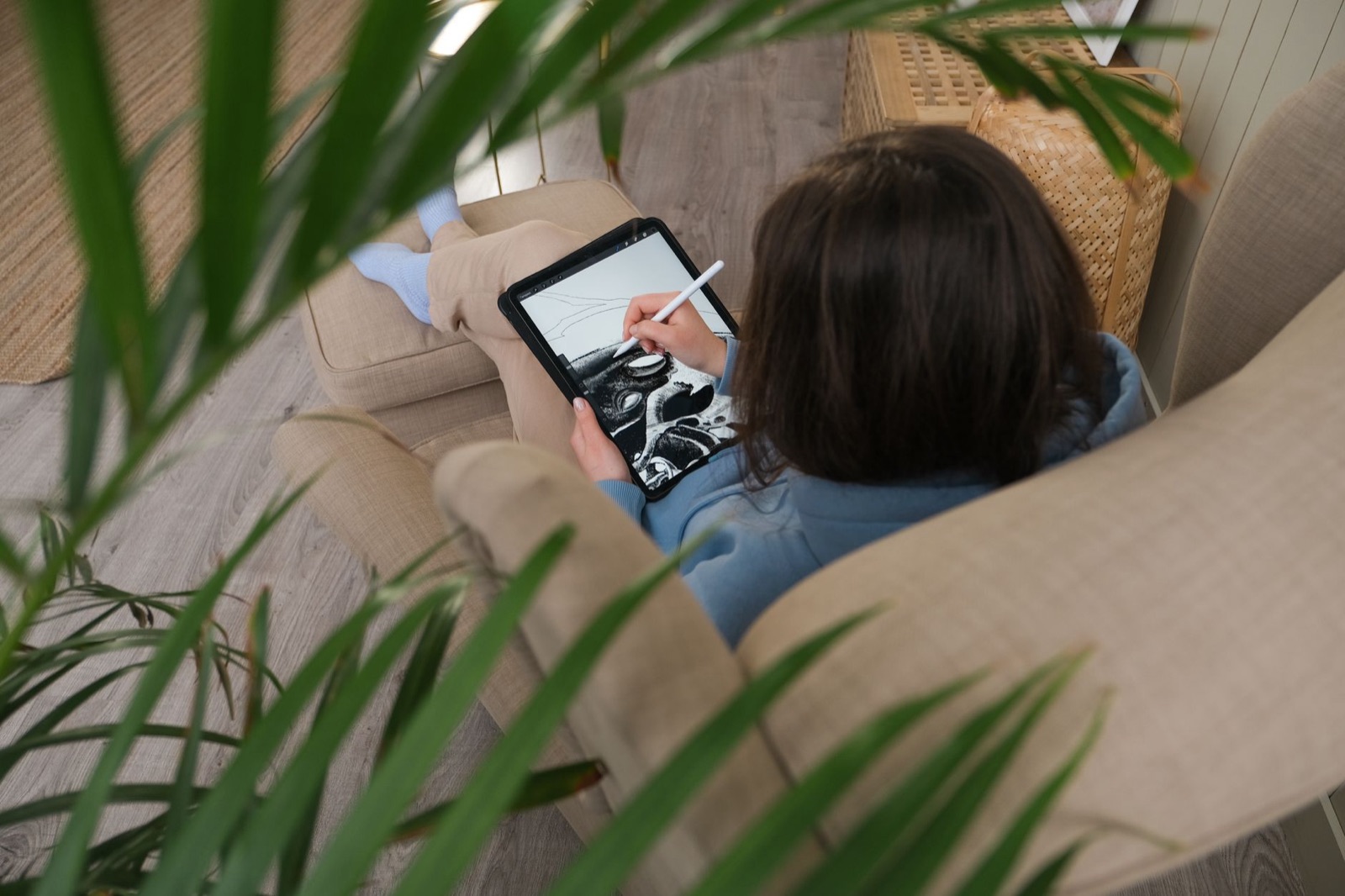 Uma pessoa desenhando em um tablet, largada em um sofá e rodeada por folhas de plantas.