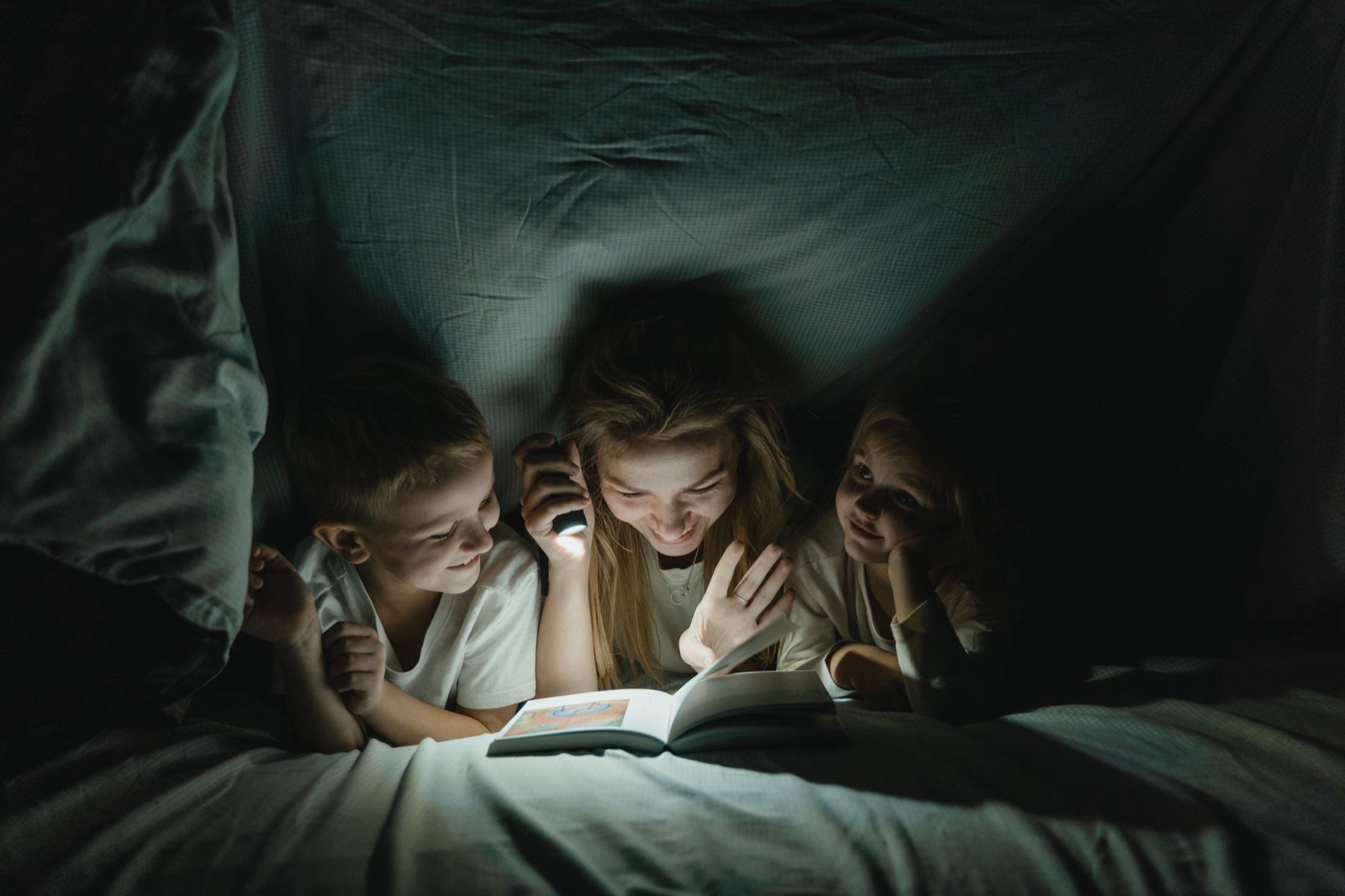 A mother shares a bedtime story with her kids under a cozy blanket, lit by a flashlight.