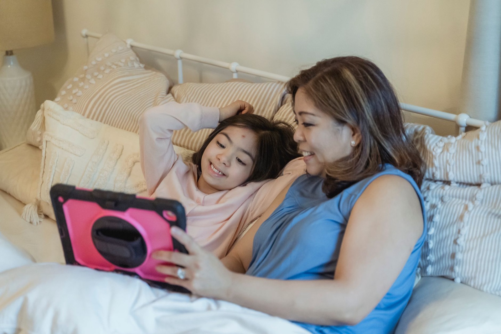 Eine Mutter und ihre Tochter lächeln, während sie zusammen auf einem Bett ein Tablet benutzen.