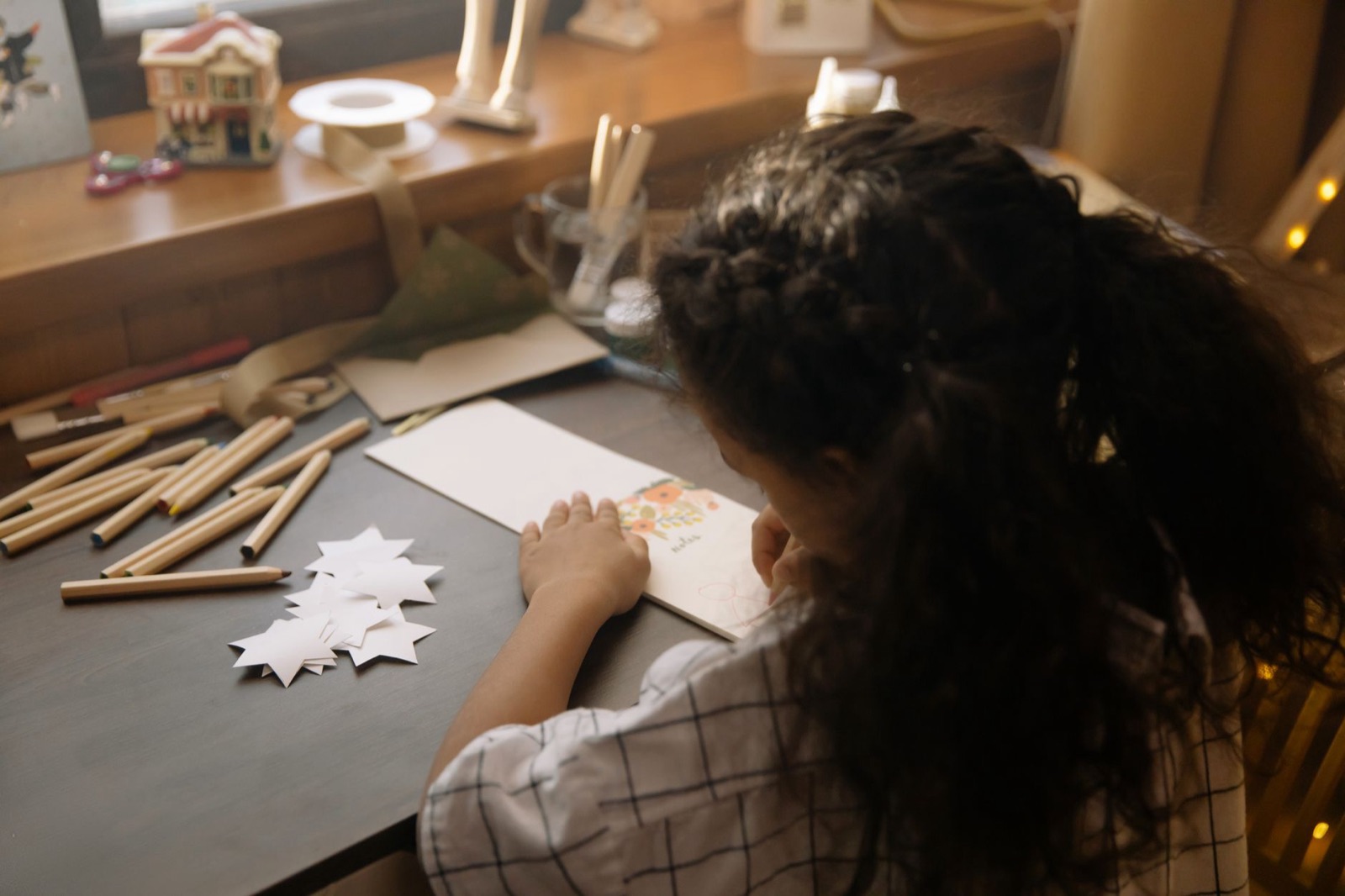 A young girl engaged in creative drawing of a Christmas card, surrounded by arts and crafts materials indoors.