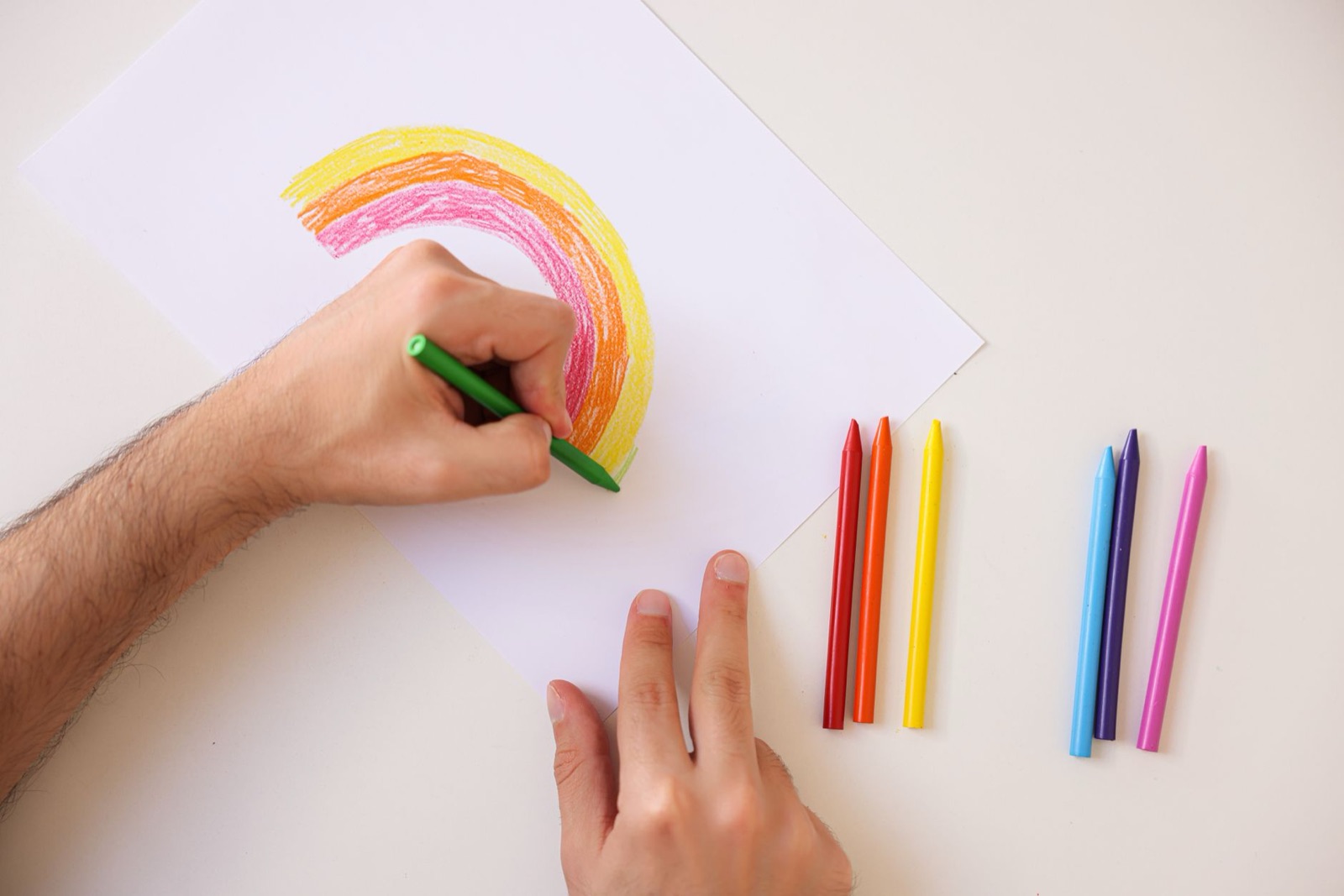 Hand drawing a colorful rainbow using crayons on white paper, showcasing creativity and art.
