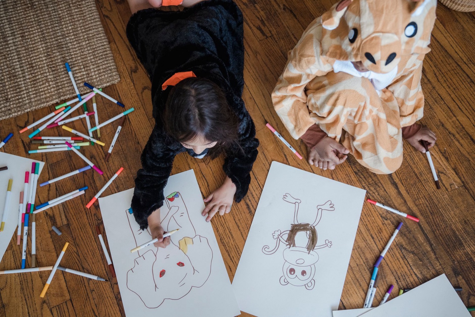 Two children in animal costumes coloring with markers on the wooden floor.