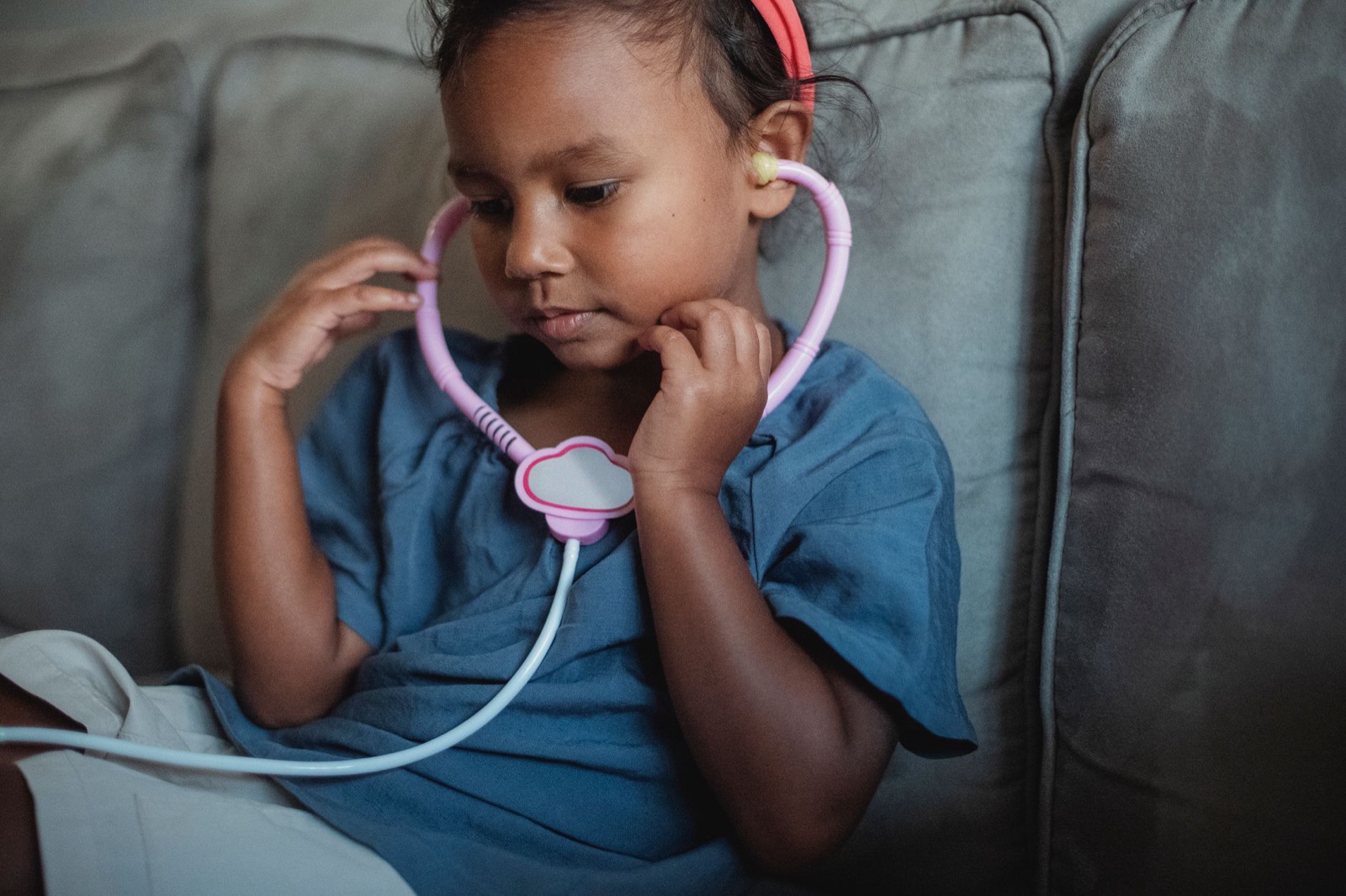 Side view of crop focused Asian kid wearing headband sitting on soft couch with toy stethoscope while playing doctor in cozy living room