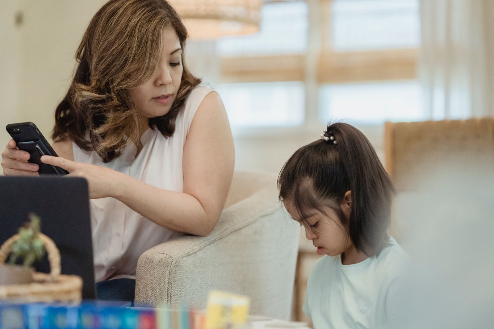 A mother using a smartphone while her daughter reads nearby, creating a warm family moment.