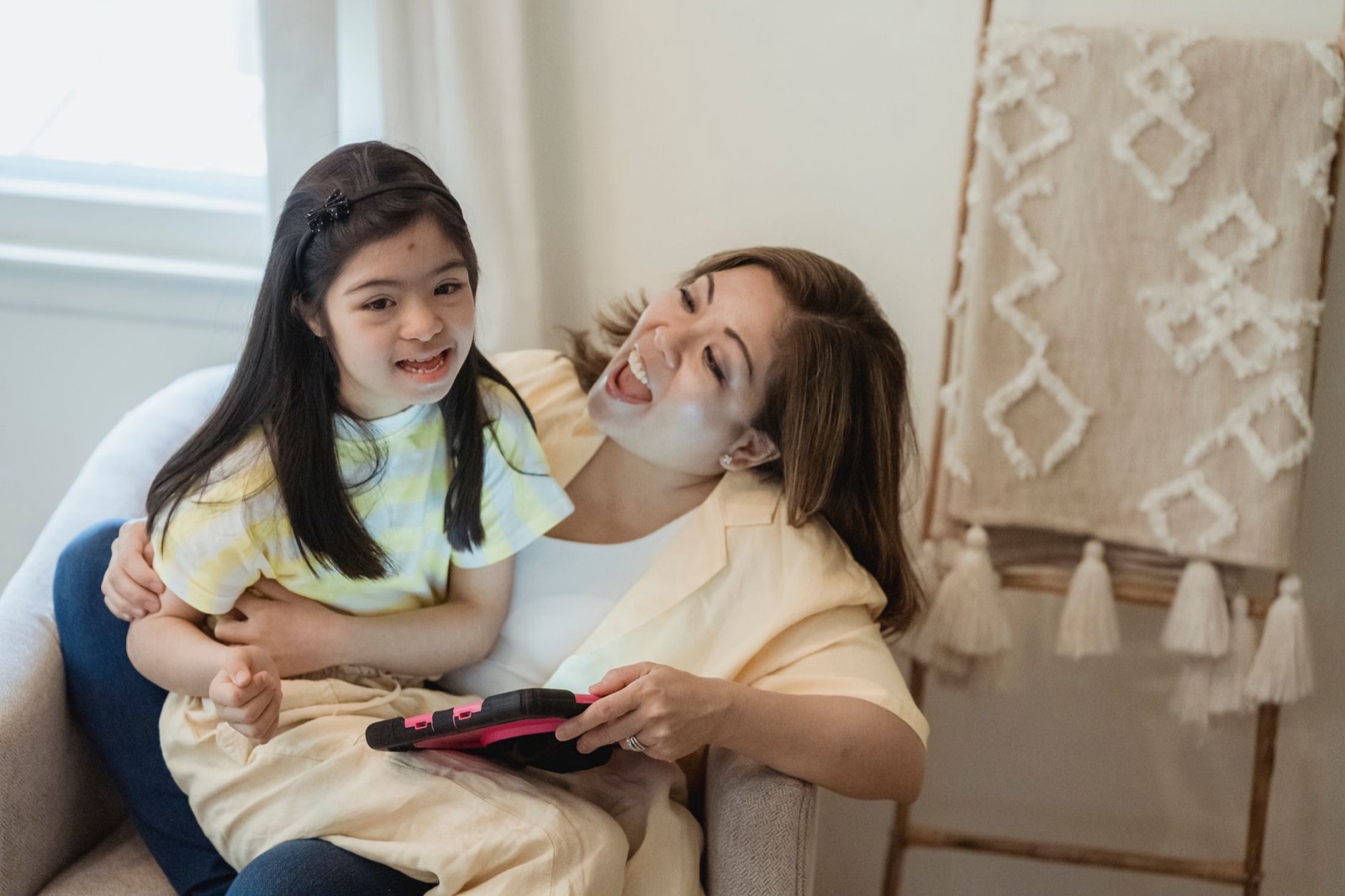 A joyful mother and daughter bonding while using a tablet, seated indoors.