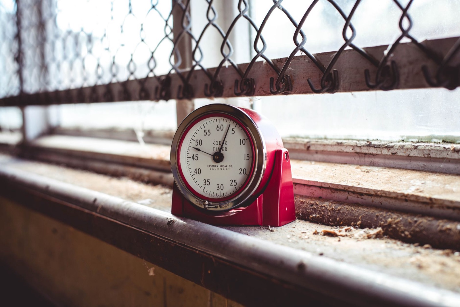 A red timer sits on a dusty windowsill with a chain link fence background, evoking nostalgia.