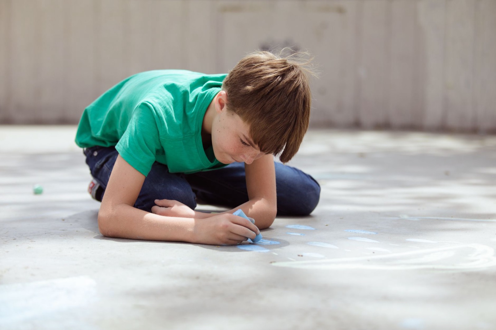 Child in a green shirt drawing on pavement with chalk during the day.