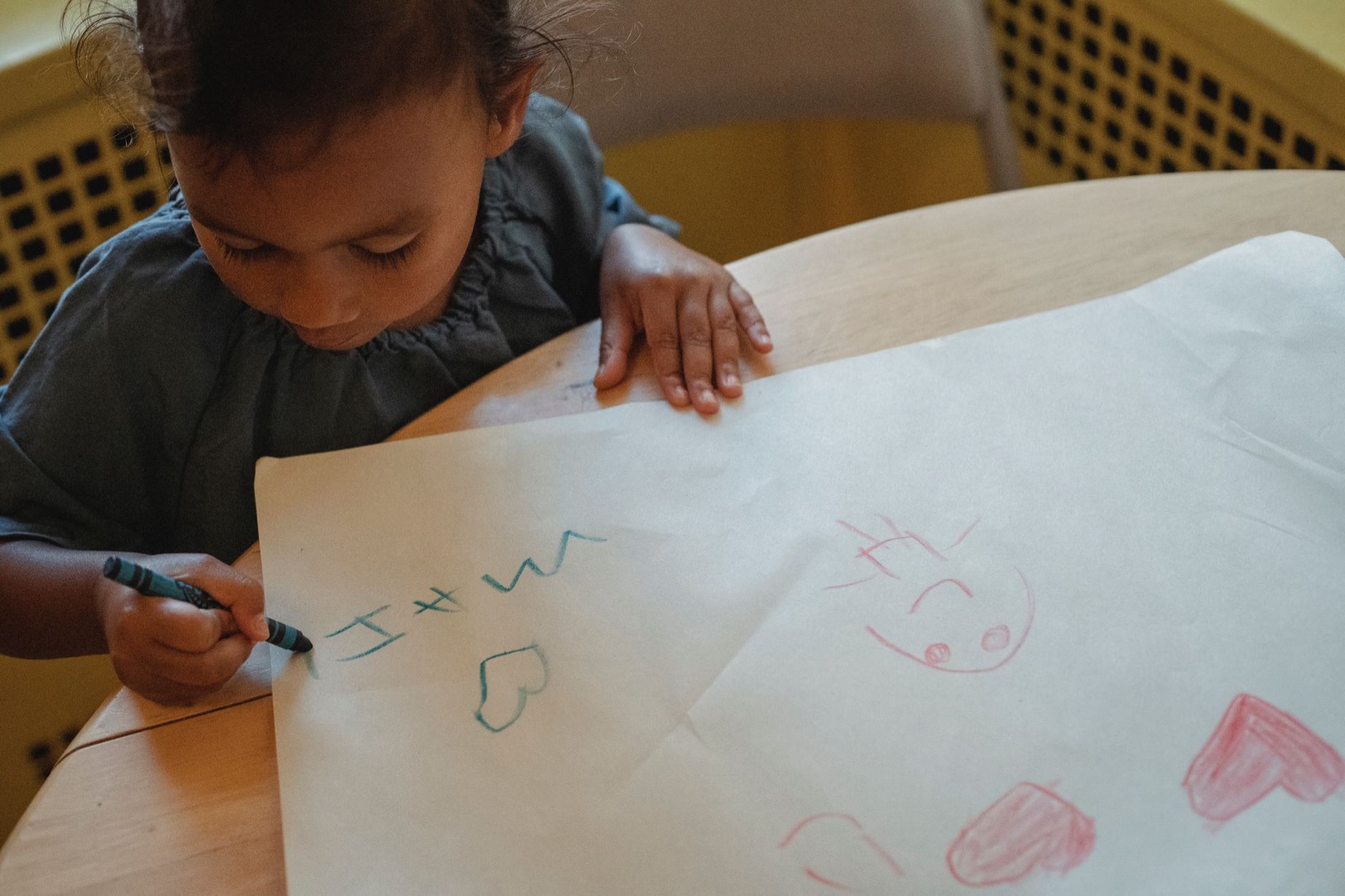 From above of concentrated little Hispanic girl drawing with wax crayons on white paper while sitting at table