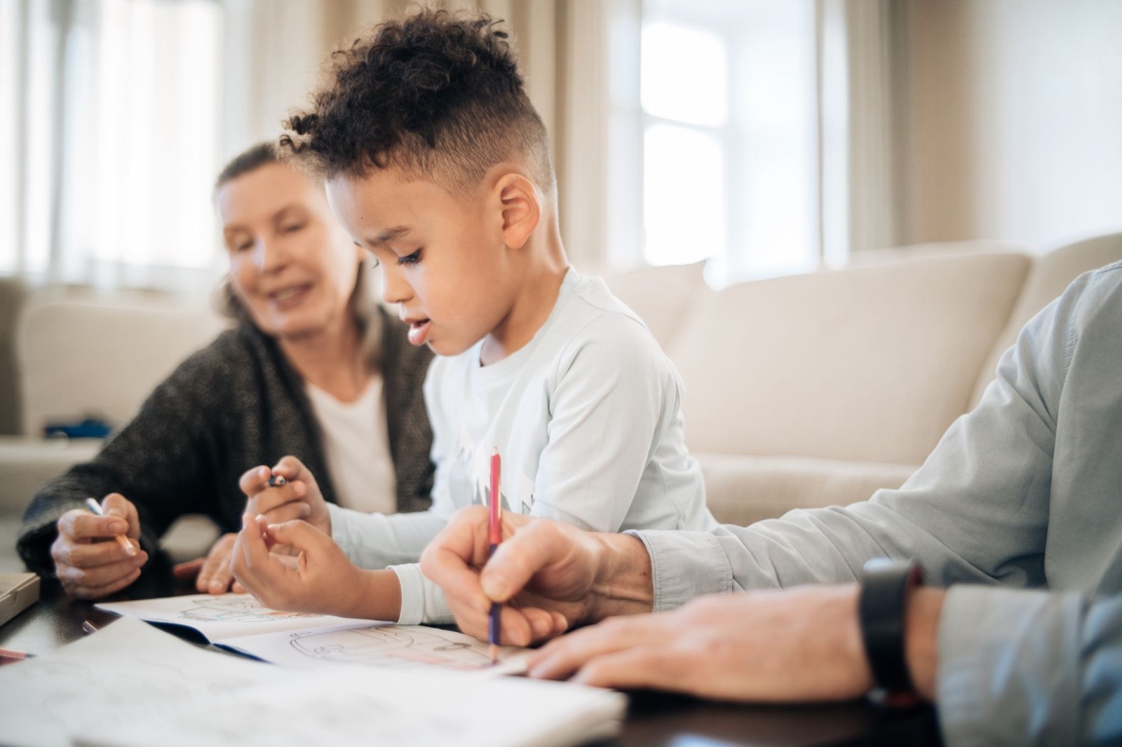 Multiracial boy with grandparents sitting at table and drawing pictures on papers with pencils in light living room near couch