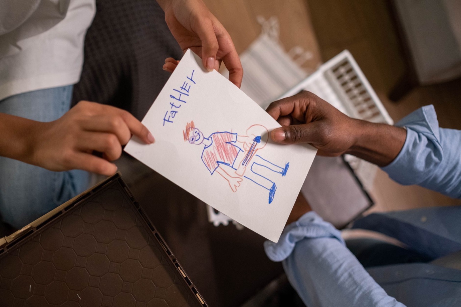 A child hands a drawing labeled 'Father' to an adult indoors, showcasing family bonding and creativity.